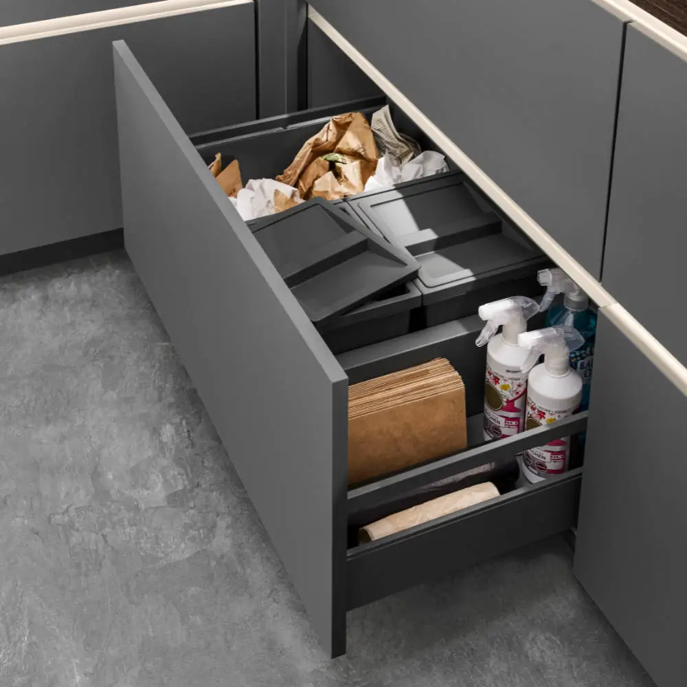 Offene graue Küchenschublade mit mehreren Fächern, die Papiertüten, Plastikbehälter mit Deckeln, Sprühflaschen und braune Papiertüten für Lebensmittel oder Mülltrennung enthält.