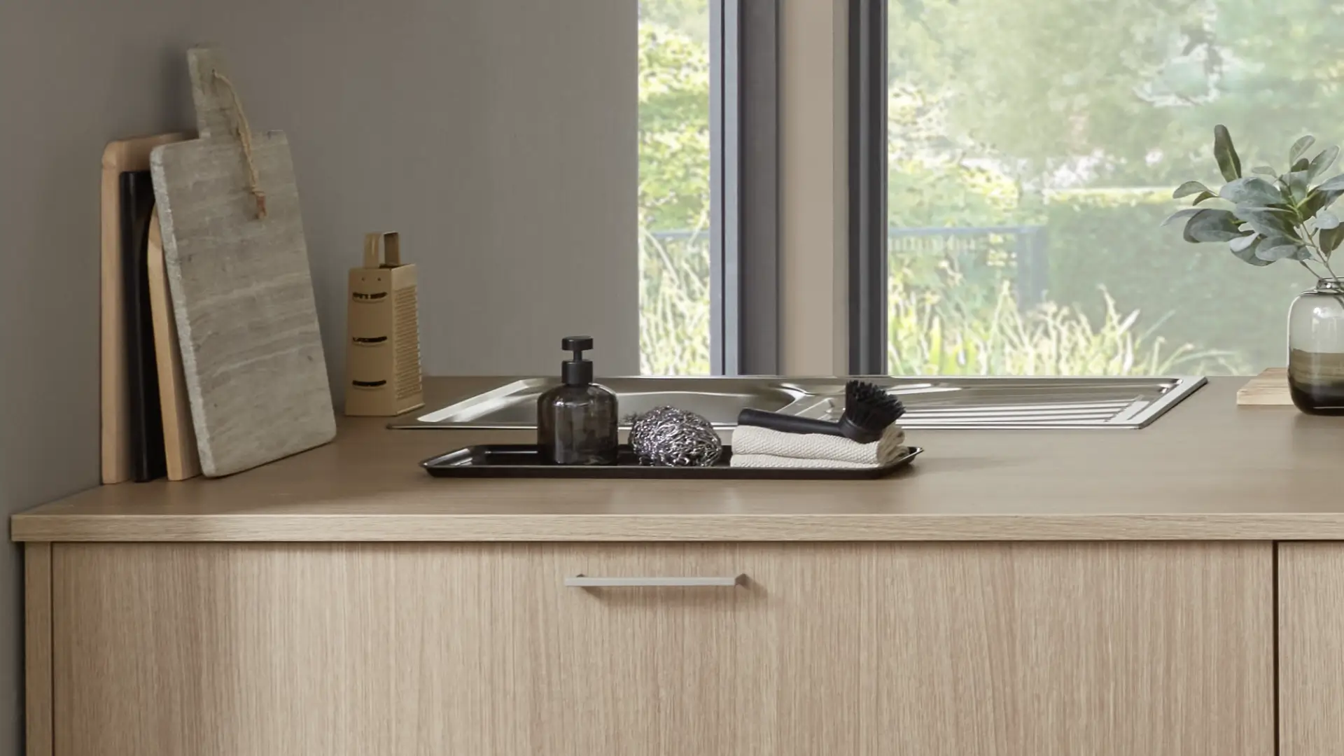 Moderner Küchenarbeitsbereich mit Holzarbeitsplatte, Edelstahlspüle, mehreren Schneidebrettern, Seifenspender, Stahlwolle, Bürste, weißen Lappen und Vase mit grünen Blättern neben großem Fenster mit Blick auf Garten.