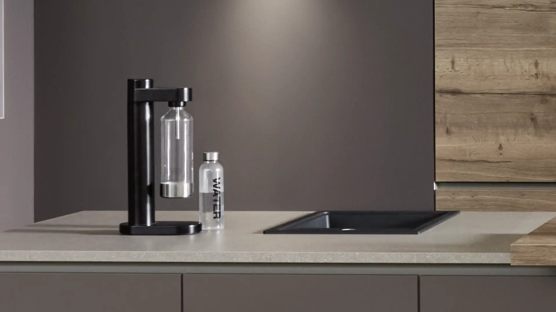 Moderne Küchenarbeitsplatte mit einem schwarzen Wassersprudler und einer transparenten Flasche mit 'WATER' Schriftzug neben einer schwarzen Spüle, im Hintergrund graue und holzfarbene Küchenschränke.