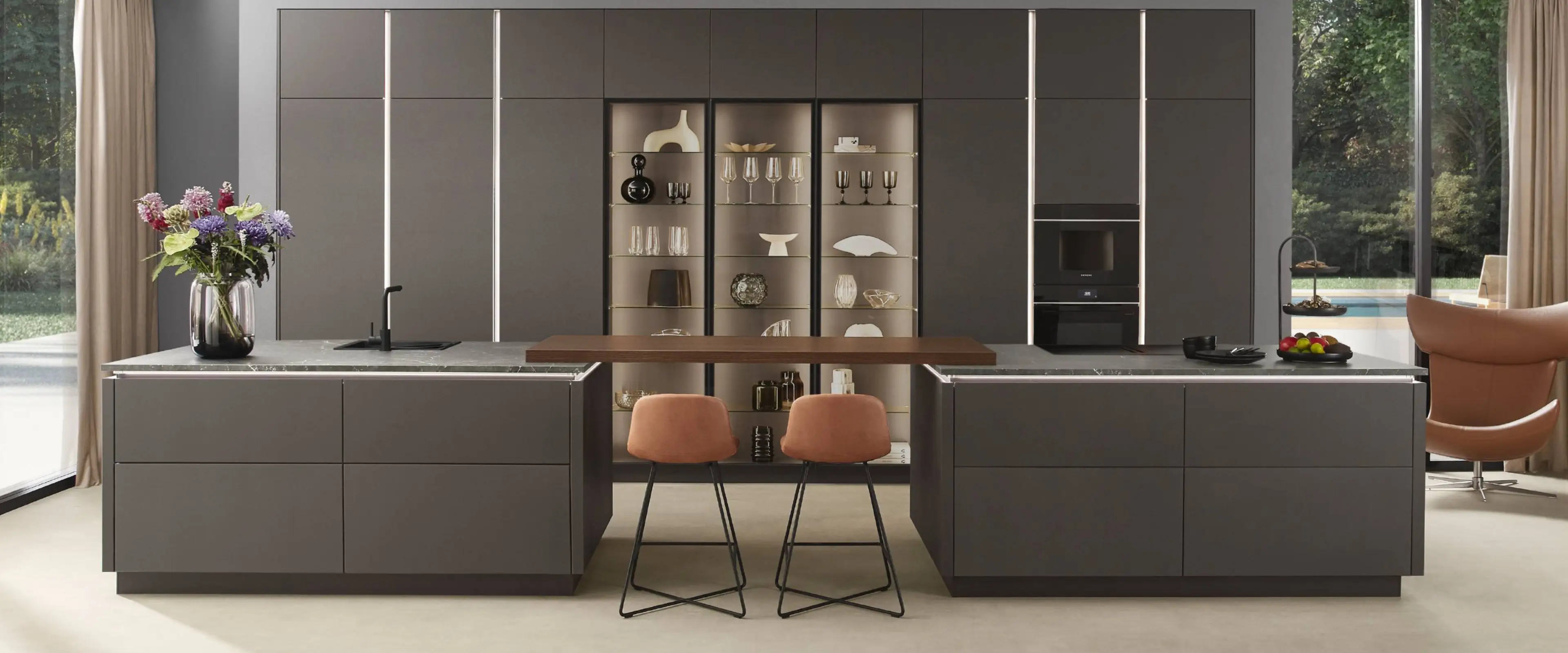 Moderne grifflose Küche mit zwei großen grauen Inseln, marmorierten Arbeitsplatten und zwei orangefarbenen Barhockern an einer Holzplatte in der Mitte, im Hintergrund Wand mit eingebauten Vitrinen, Gläsern und Dekoration, seitlich große Fenster mit Blick auf Garten und Terrasse.