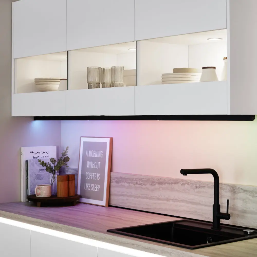 Moderne Küchenzeile mit weißem Oberschrank mit beleuchtetem Glasfach, darunter Arbeitsplatte mit schwarzer Spüle und mattschwarzem Wasserhahn, dekoriert mit Bildern, Keramikbechern und Pflanzen, sanfte Farbbeleuchtung an der Wand.