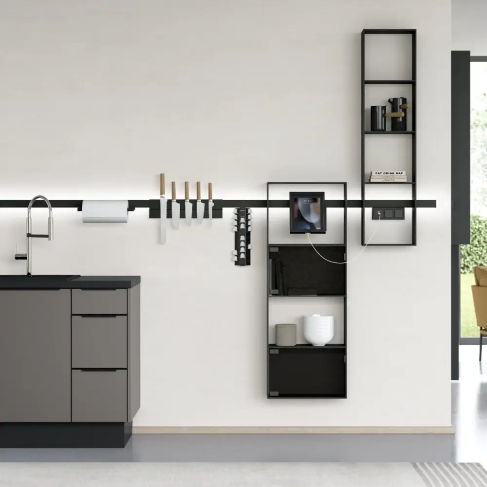 Moderne Küchenwand mit schwarzem Relingsystem, das Messer, Papierrollenhalter, Tassenspender, Gewürzregal und zwei offene Regale hält, daneben graue Küchenunterschränke mit Wasserhahn.