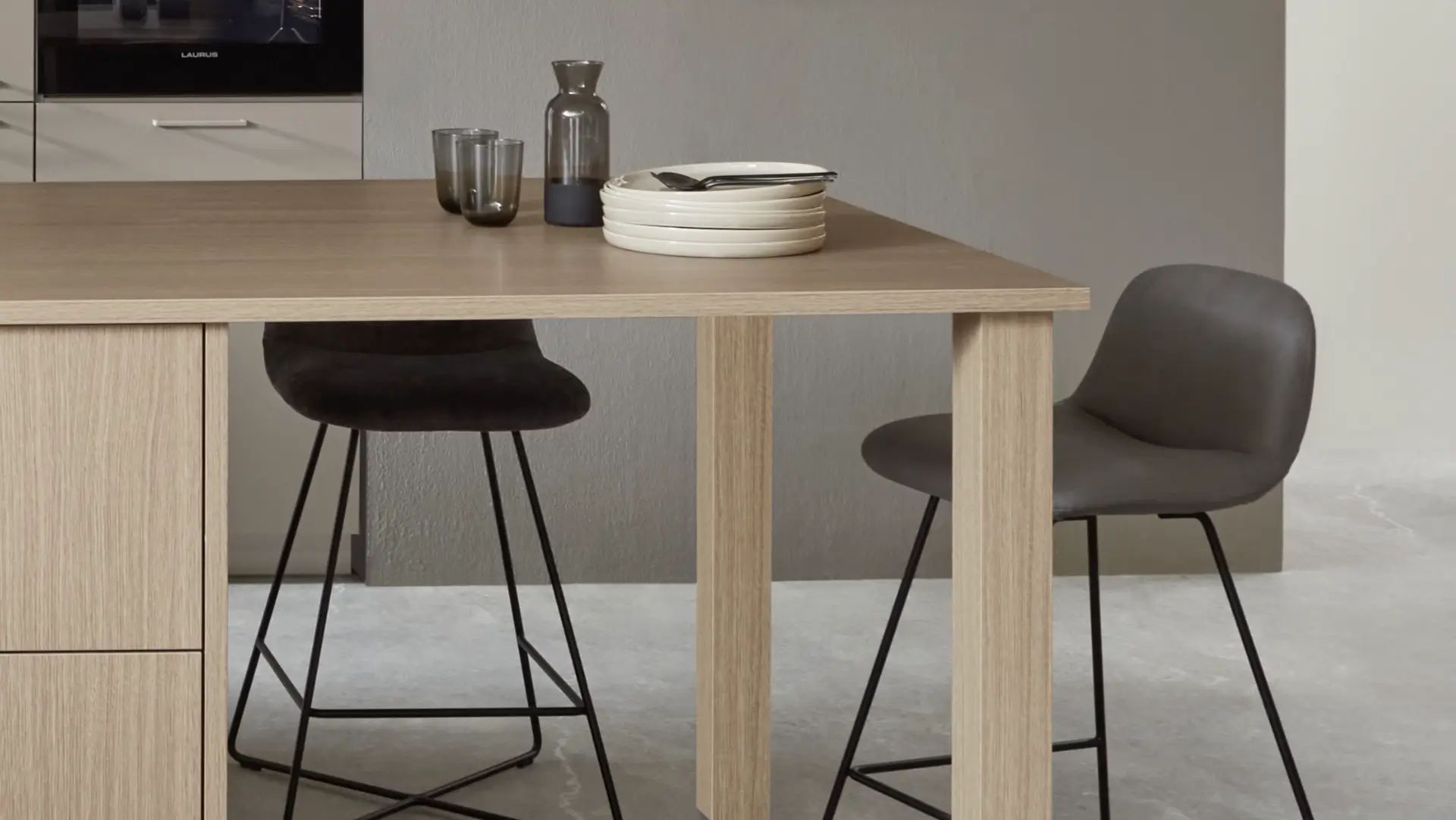 Moderner Esstisch aus hellem Holz mit schwarzen Metallstühlen, auf dem Tisch stehen weiße Teller, Besteck und zwei Gläser neben einer kleinen Karaffe.