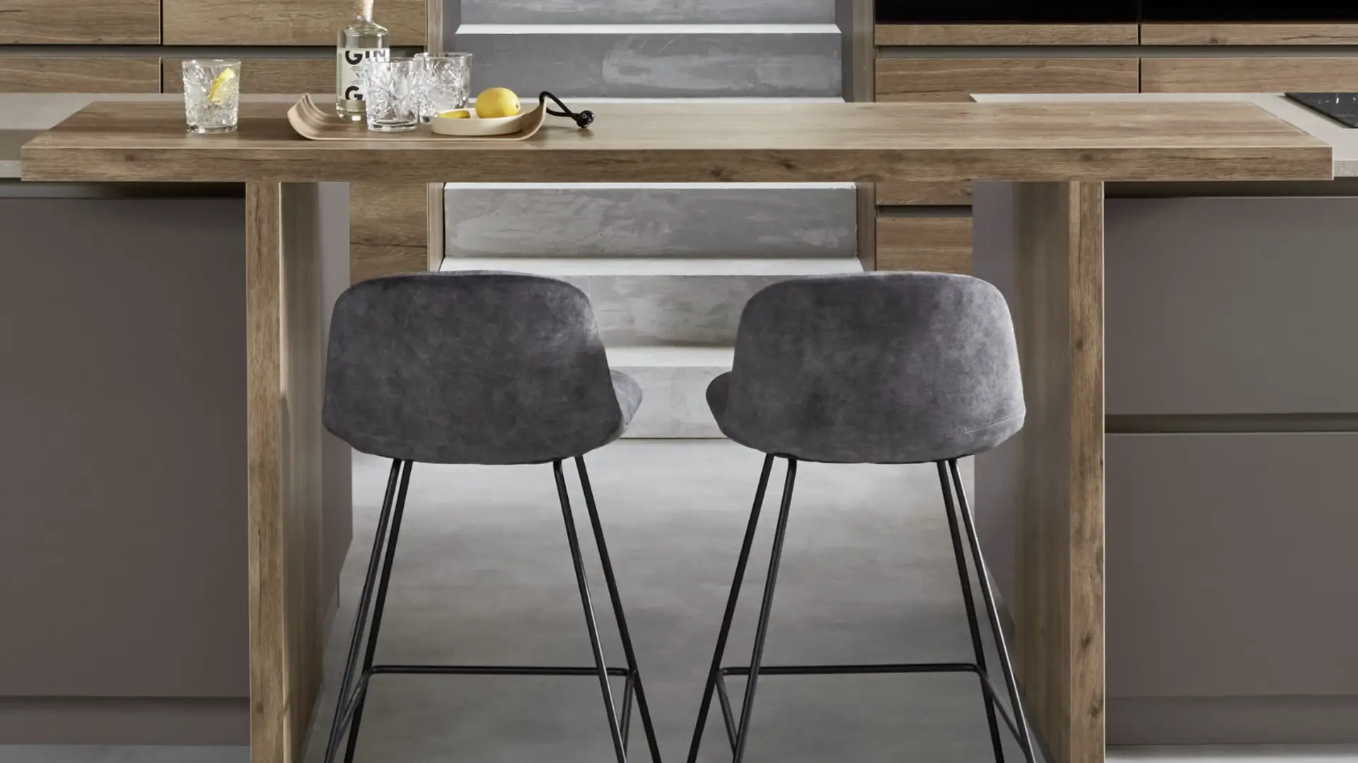 Moderne Küchentheke aus hellem Holz mit zwei grauen Barhockern mit Metallbeinen, Gläsern und Zitronen auf der Arbeitsplatte, im Hintergrund Sitzstufen aus Beton.