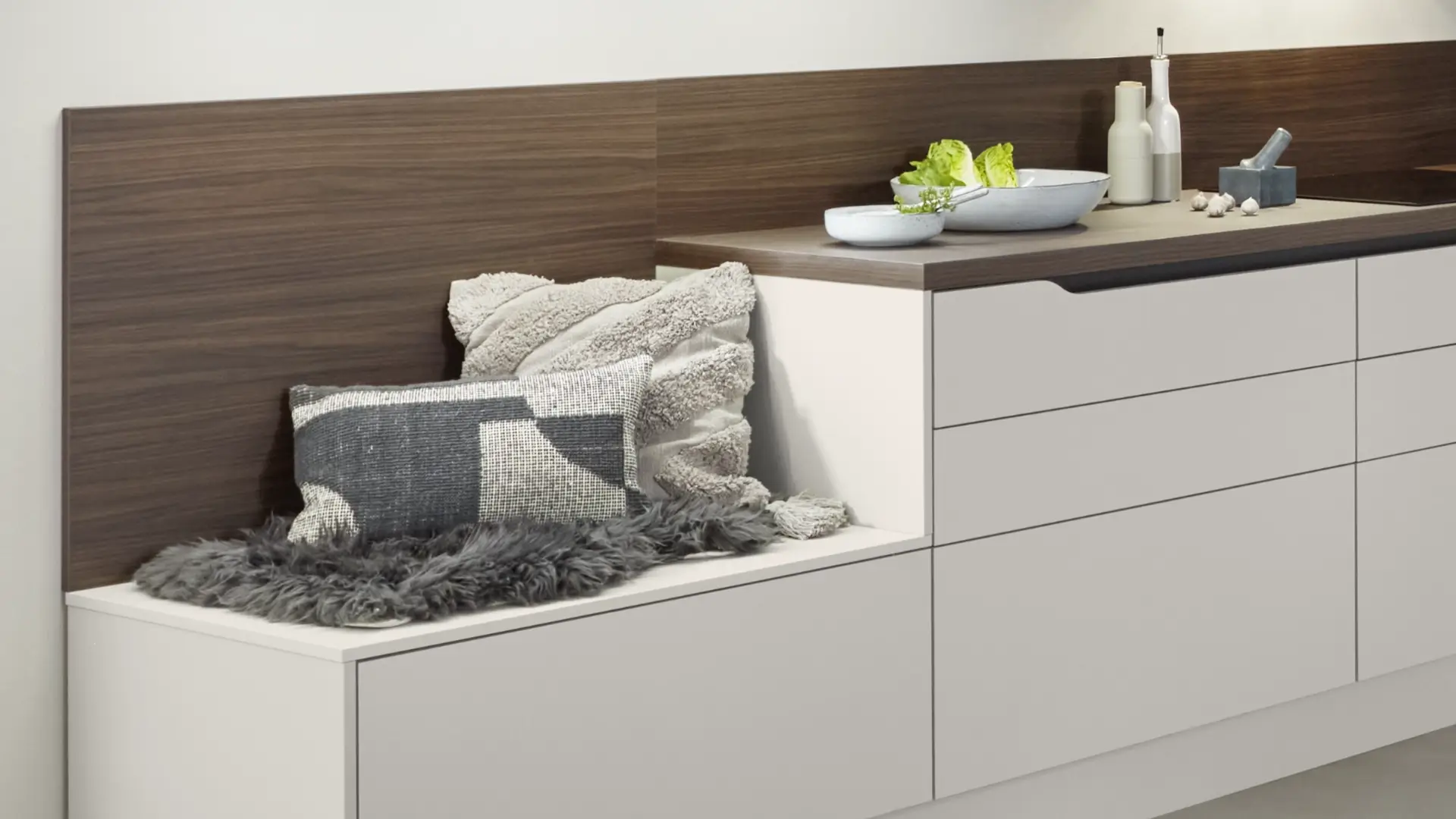 Moderne Eckküche mit weißen Schränken und Holzarbeitsplatte, Sitzecke mit grauem Fell und zwei dekorativen Kissen, auf der Arbeitsplatte Schalen mit grünem Salat, Gewürzflaschen und Knoblauch.