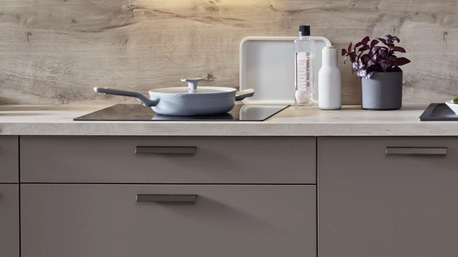 Moderner Küchenarbeitsbereich mit grauen Schubladen, einem grauen Topf mit Deckel auf einer schwarzen Induktionskochplatte, einem weißen Tablett, einer Glasflasche mit rosa Himalaya-Salz, einer weißen Flasche und einem Topf mit dunklen Pflanzen auf einer hellen Holzplatte und Holzrückwand.