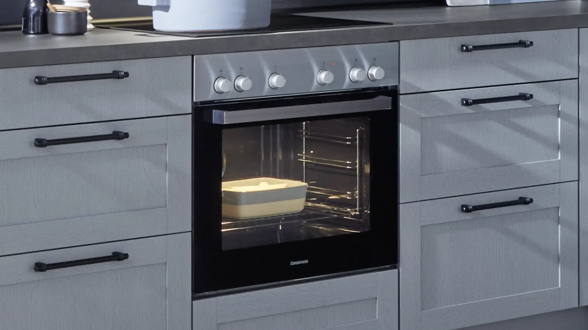 Moderne graue Küchenschränke mit schwarzen Griffen und eingebautem Backofen, in dem eine Auflaufform mit Essen steht.