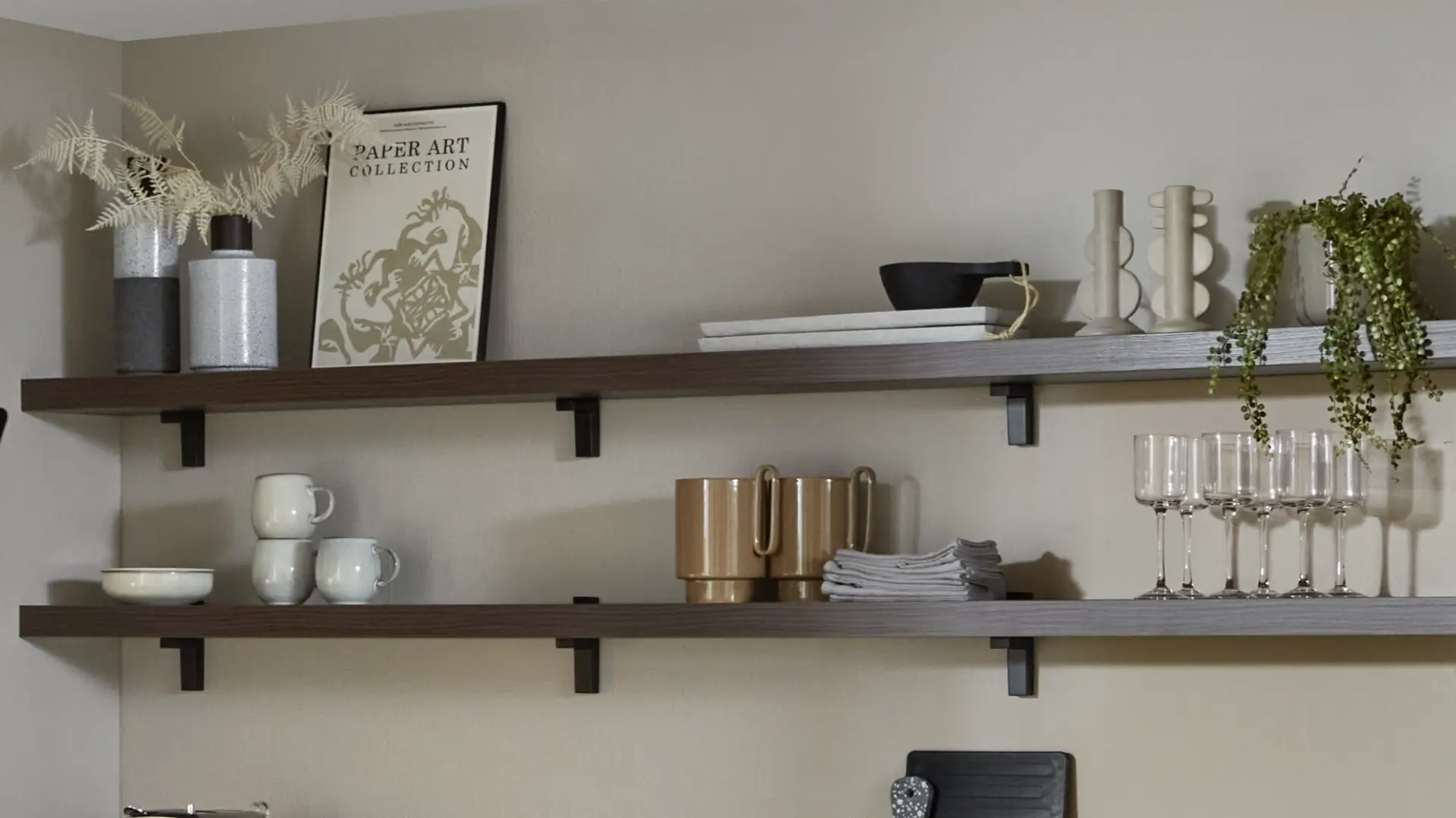 Zwei hölzerne Wandregale mit Dekoration: Oben zwei Vasen mit getrockneten Blättern, ein gerahmtes Bild mit der Aufschrift 'PAPER ART COLLECTION', zwei Kerzenständer, eine kleine Schale und eine hängende grüne Pflanze; unten eine weiße Schale, drei weiße Tassen, zwei beige Keramikbecher, ein Stapel grauer Servietten und fünf Weingläser.