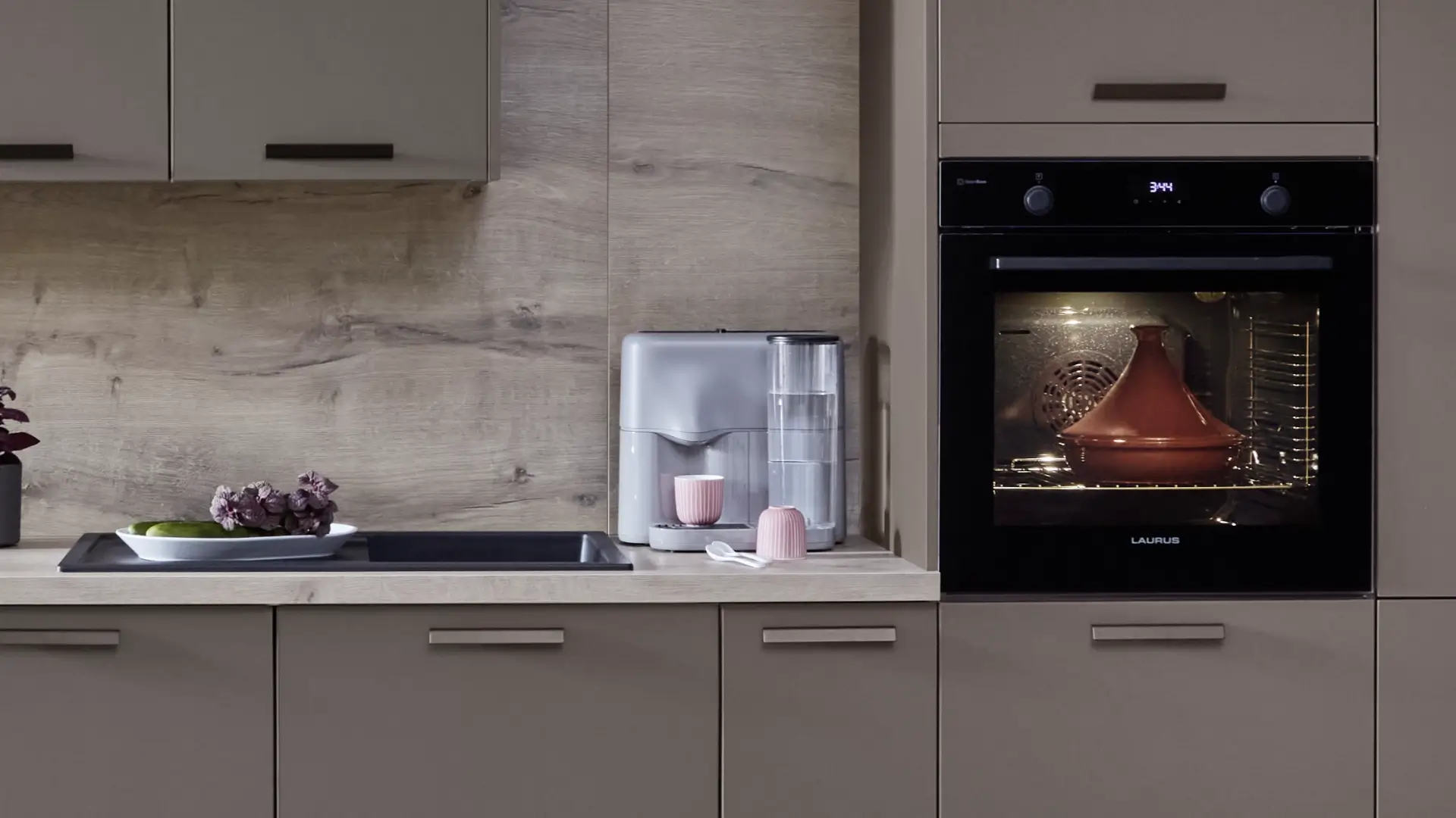 Moderne Küchenzeile mit grauen Schränken, einem eingebauten Ofen, in dem ein Keramiktopf steht, einer Kaffeemaschine mit zwei rosa Bechern und einer weißen Schale mit Gemüse auf der Arbeitsplatte vor hellem Holzrückwand.