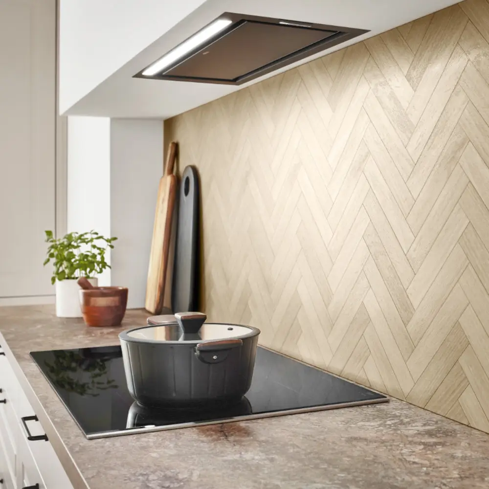 Moderne Küche mit eingebautem Induktionskochfeld, auf dem ein schwarzer Topf mit Glasdeckel steht, eine Küchenarbeitsplatte in Steinoptik, Holz-Schneidebretter und ein Kräutertopf im Hintergrund, sowie eine wandmontierte Dunstabzugshaube über dem Kochfeld.