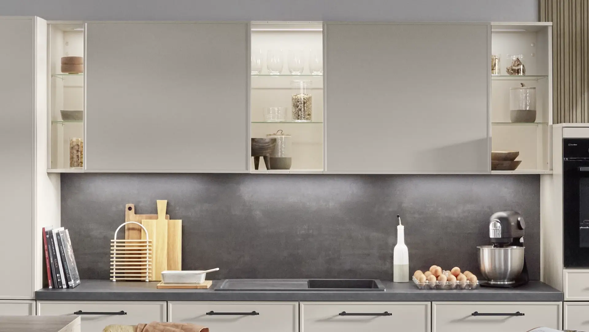 Moderne Küche mit hellen Oberschränken, in denen Gläser und Vorratsgläser sichtbar sind, eine Arbeitsplatte mit einem Spülbecken, Schneidebrettern, einem aromatisierten Ölspender, einem Eierkarton und einer Küchenmaschine vor einer dunklen Rückwand.