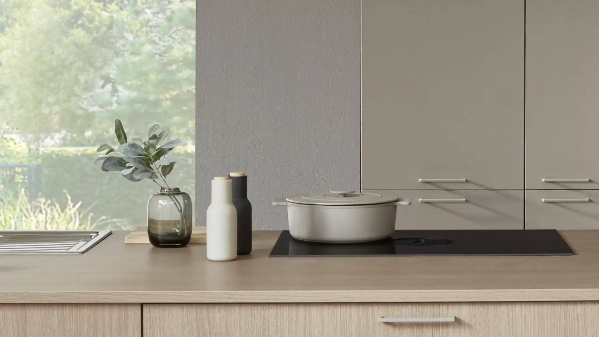 Moderne Küchenarbeitsplatte aus hellem Holz mit einem schwarzen Kochfeld, darauf ein grauer Topf mit Deckel, daneben zwei kleine Flaschen in Schwarz und Weiß und eine Vase mit grünen Zweigen, im Hintergrund graue Küchenschränke und ein Fenster mit Blick auf grüne Pflanzen.