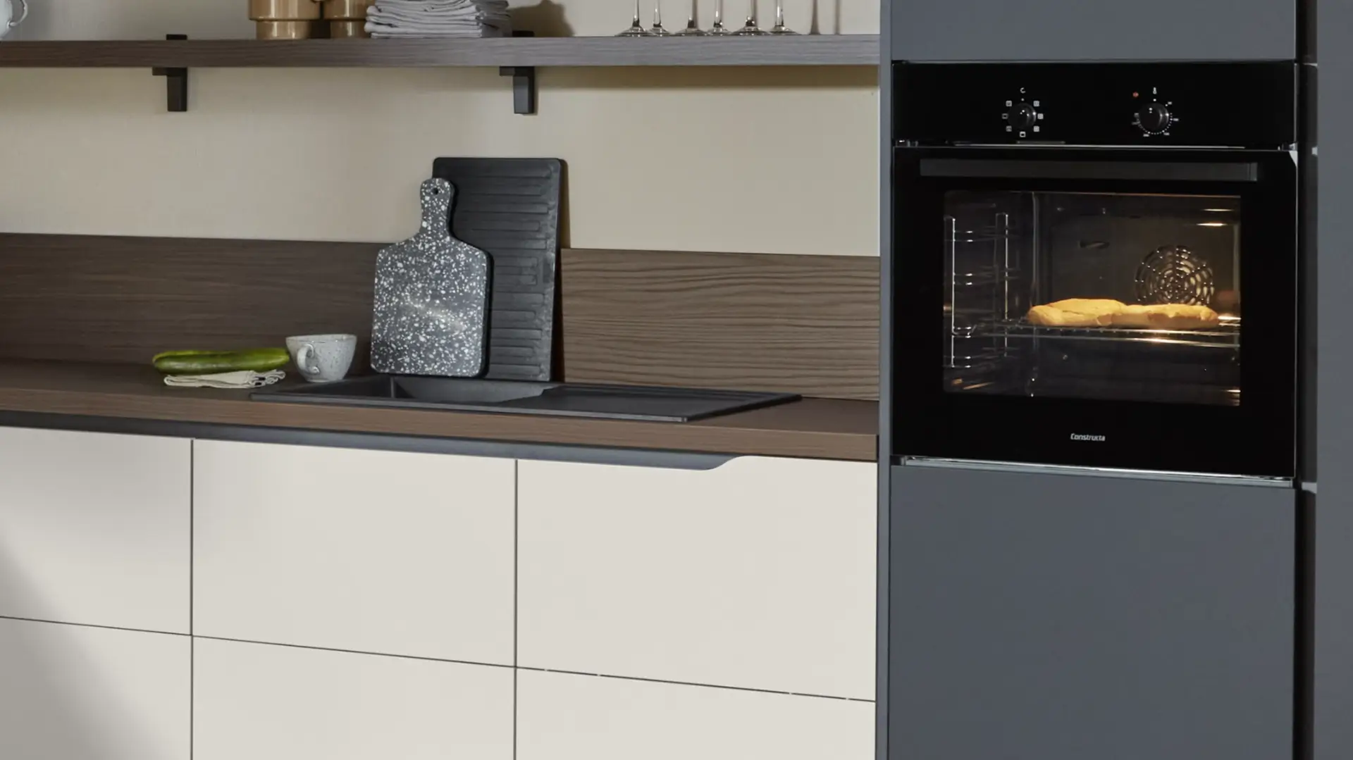 Moderne Küche mit hellen Schrankfronten, einer Arbeitsplatte mit Spüle, Holzrückwand, schwarzen Schneidebrettern und einem eingebauten Ofen, in dem Brot backt.
