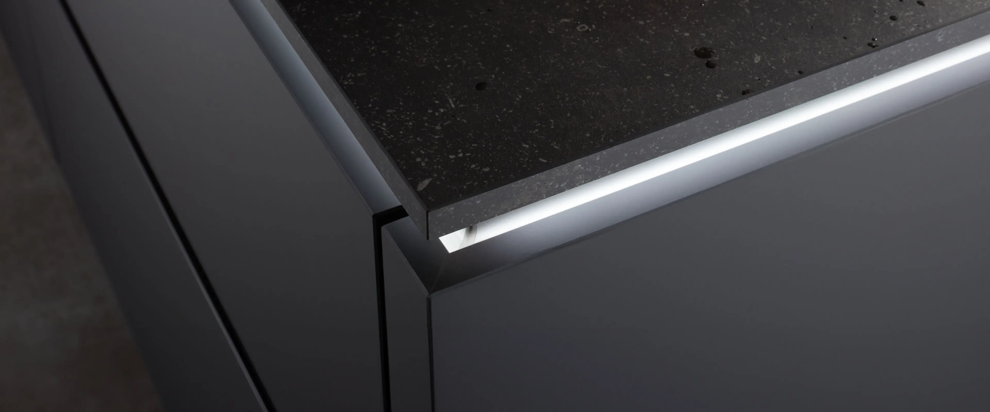 Nahaufnahme einer modernen Küchenecke mit mattschwarzen Schränken und einer schwarzen Küchenarbeitsplatte mit subtiler Textur und weißen LED-Lichtstreifen unter der Arbeitsplatte.