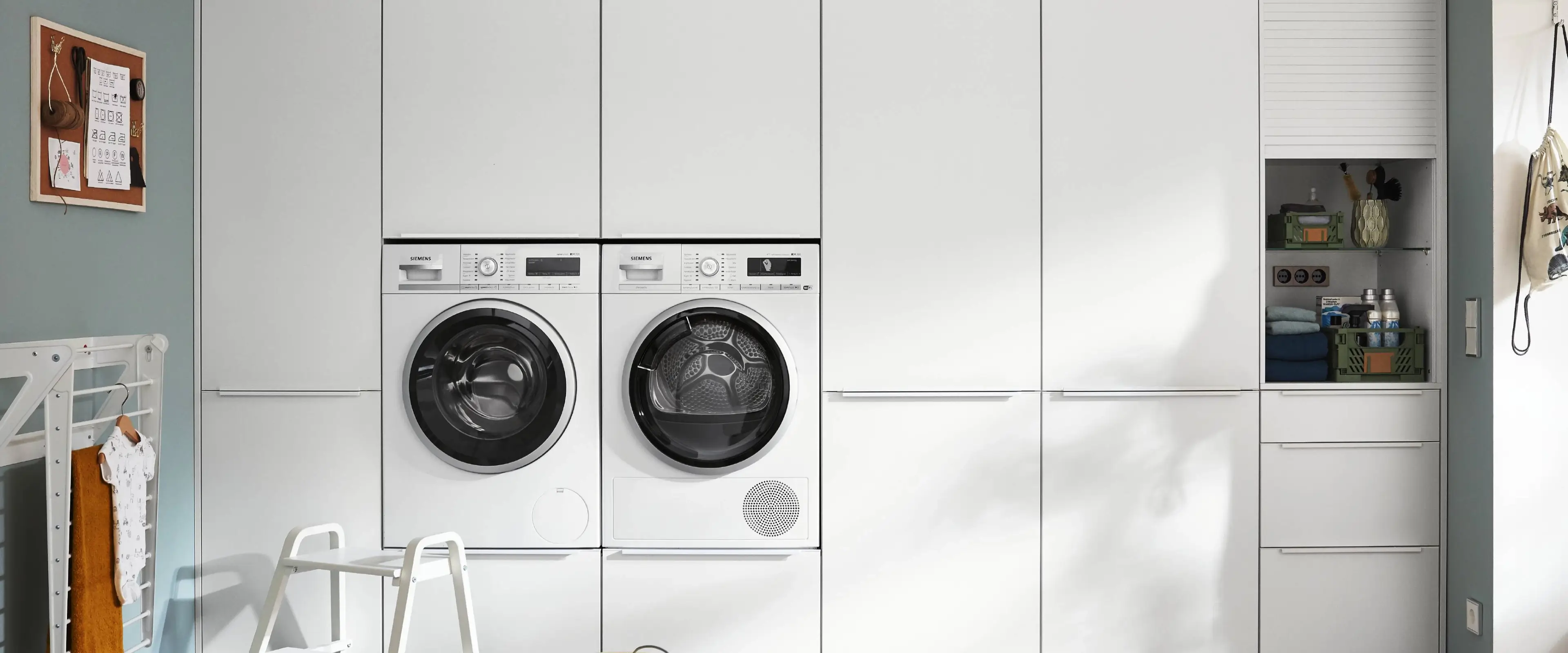 Moderner Hauswirtschaftsraum mit zwei nebeneinander eingebauten weißen Siemens-Waschmaschinen in einer weißen Einbauschrankwand, geöffnetes Fach mit gefalteten Handtüchern und Haushaltsutensilien, blau gestrichene Wand mit Pinnwand und Wäscheständer mit Babystrampler.