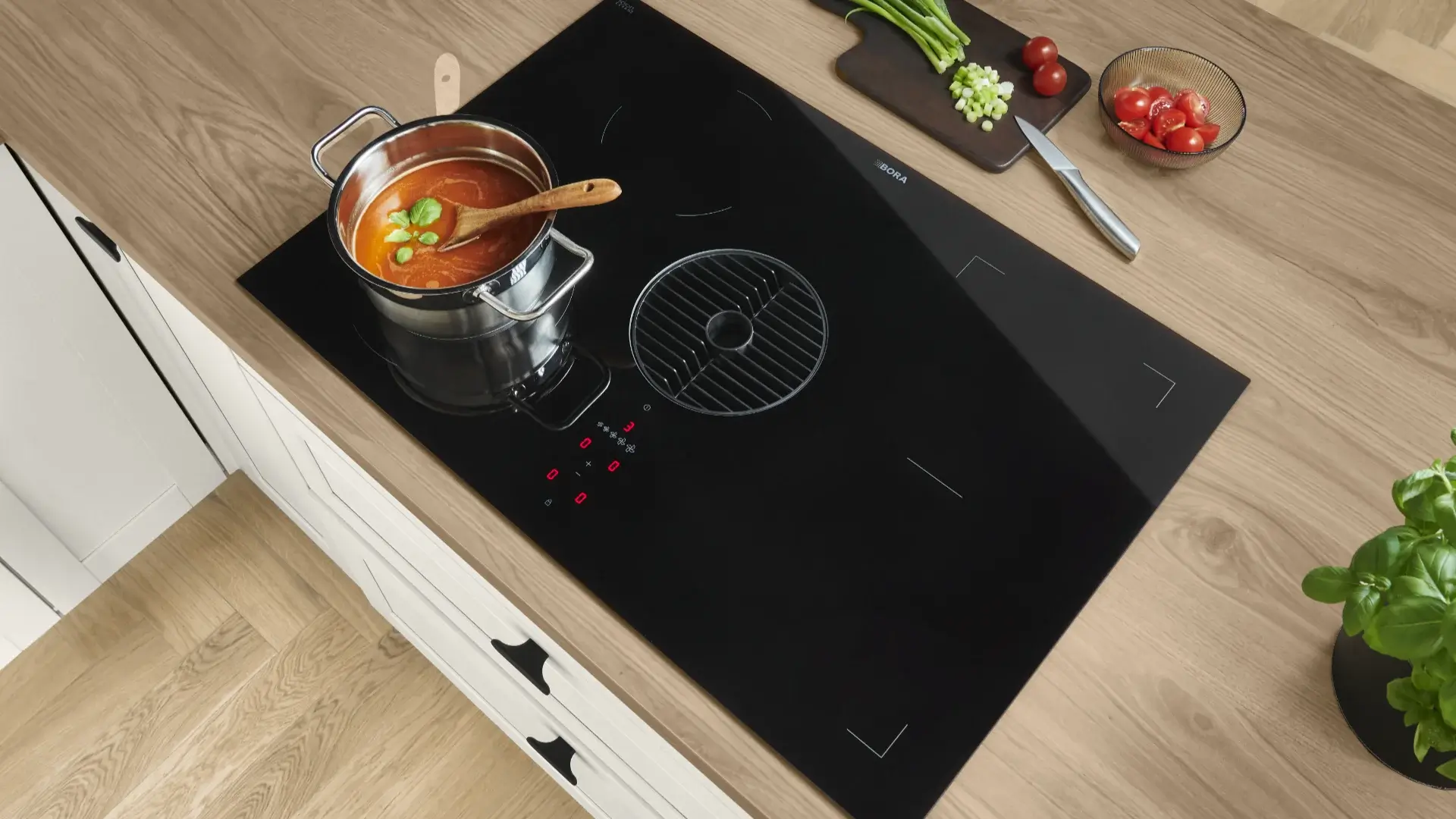 Moderne schwarze Induktionskochfeld mit integriertem Dunstabzug in einer hellen Holzarbeitsplatte, darauf ein Topf mit roter Suppe und Basilikum, daneben Schneidebrett mit geschnittenen Frühlingszwiebeln und Tomaten sowie ein Messer.