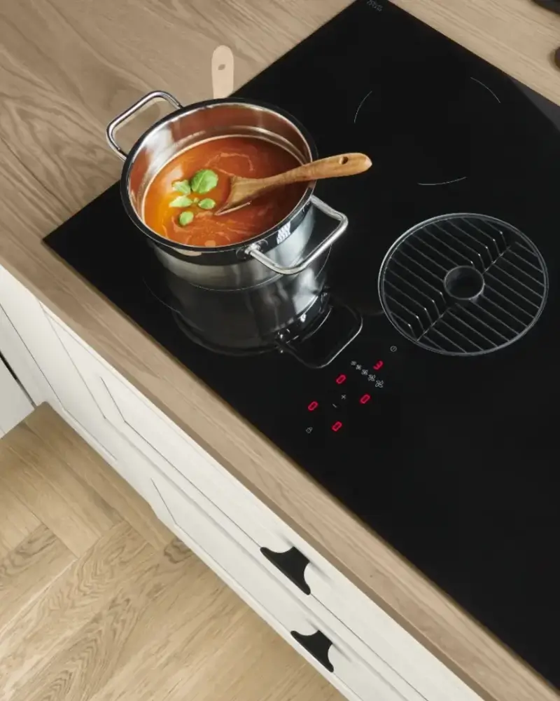 Ein Kochtopf mit Tomatensuppe, garniert mit Basilikumblättern, steht auf einem eingeschalteten schwarzen Induktionskochfeld in einer modernen Küche mit Holzarbeitsplatte und weißen Schubladen darunter.