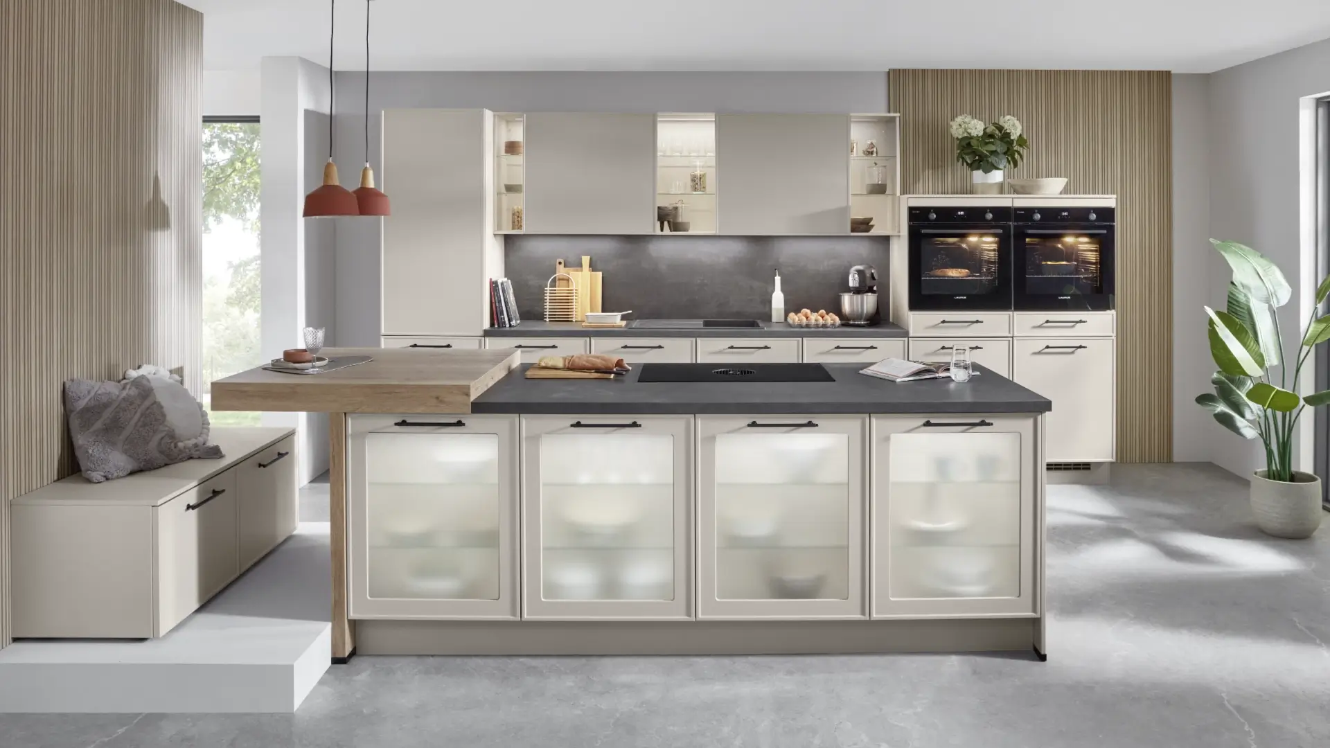 Moderne Küche mit sandfarbenen Schränken, zwei eingebauten Öfen, Kücheninsel mit dunkler Arbeitsplatte und Glastüren, Bank mit Kissen, hängenden Lampen und großen Fensterflächen.