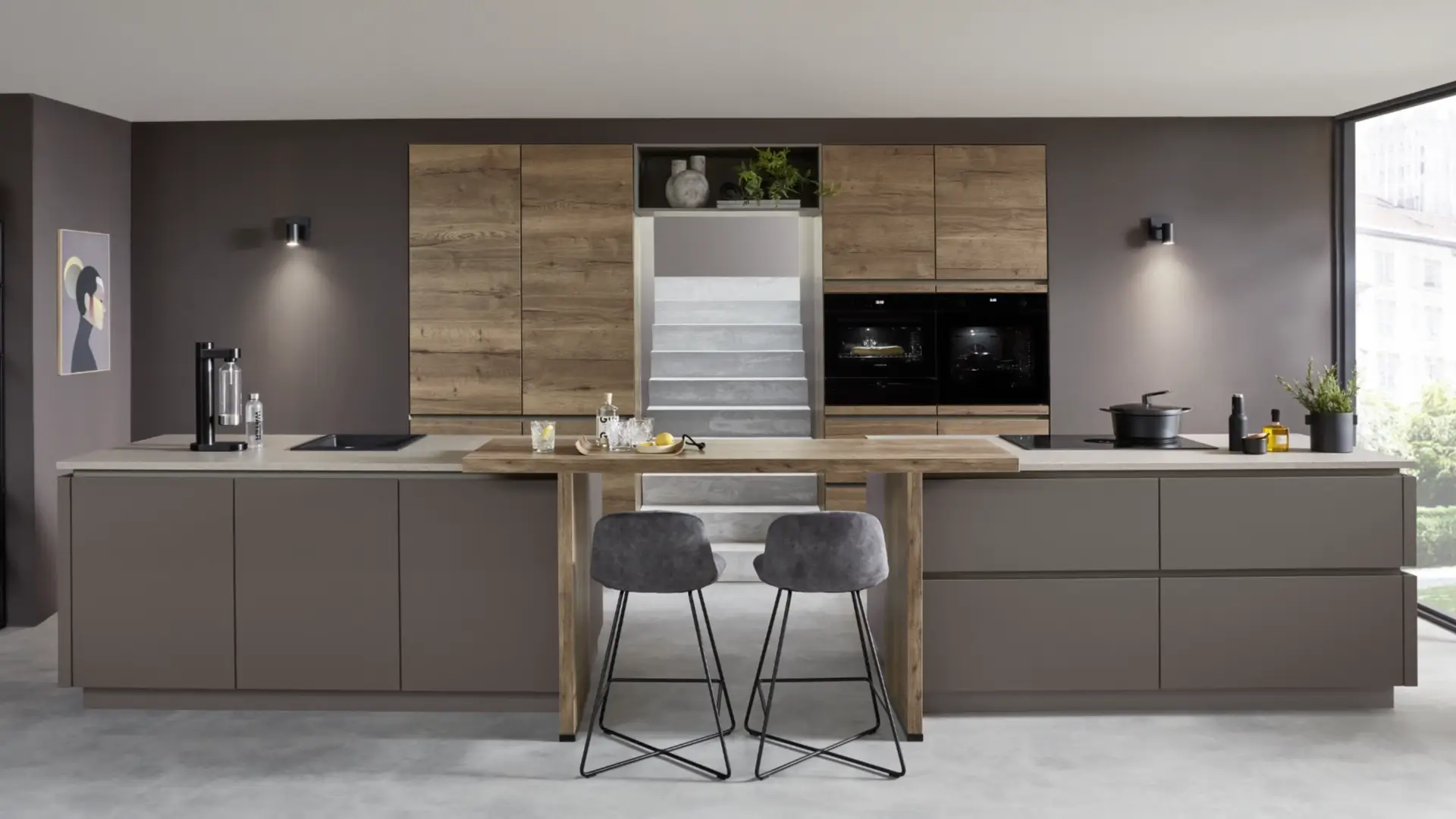 Moderne Küche mit zwei Kücheninseln: Die vordere Insel hat eine helle Arbeitsplatte mit Holz-Bar und zwei grauen Barhockern, die hintere Insel und Wandschränke sind in dunklem Grau und Holzoptik gestaltet, mit eingebautem Ofen, Kochfeld und Spüle. Die Wände sind dunkelgrau, links hängt ein Kunstbild, rechts ein großes Fenster mit Blick ins Grüne.