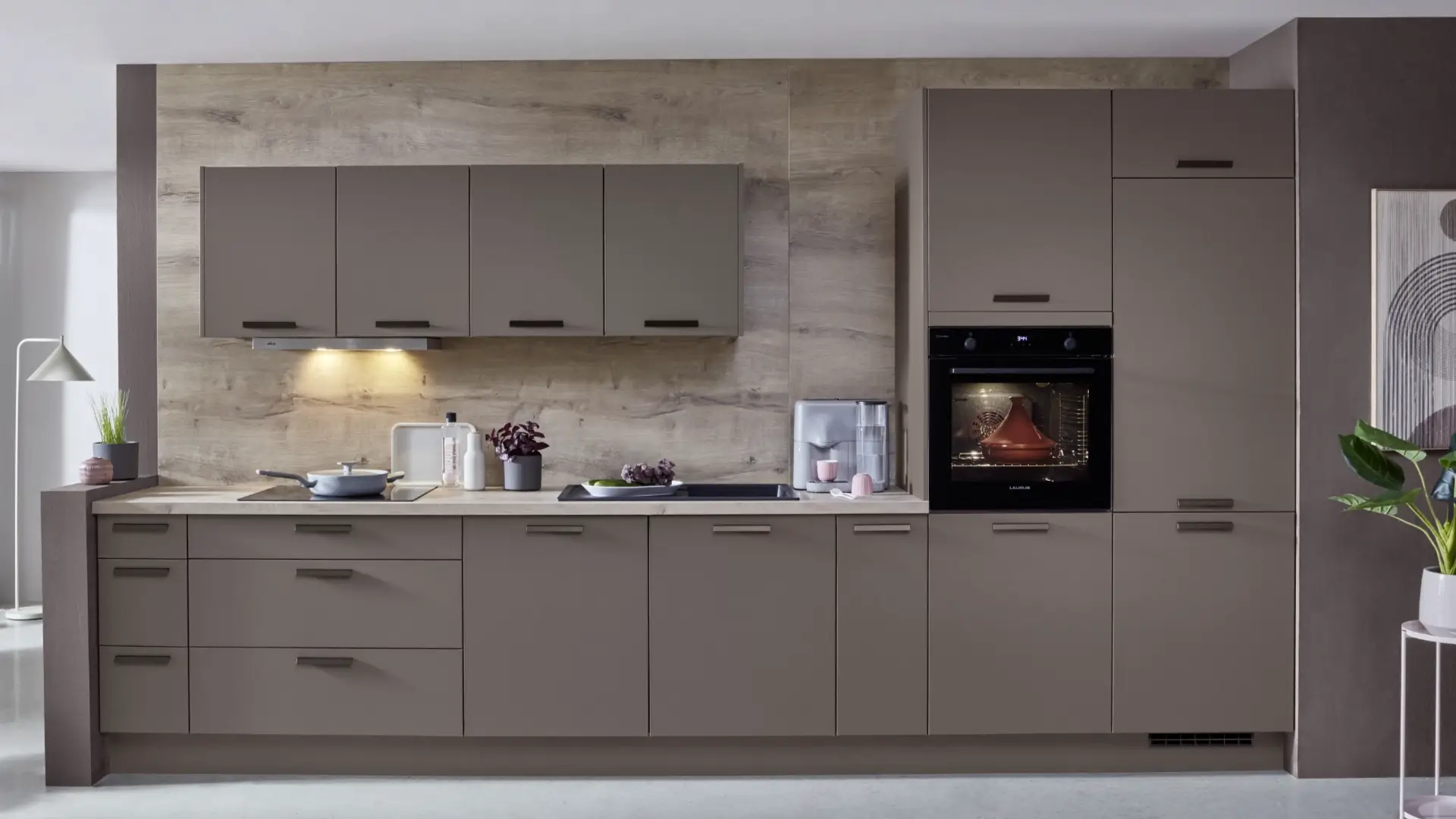 Moderne Küche mit matten, graubraunen Schrankfronten und Griffen, integrierter Backofen mit eingeschaltetem Licht, Kochfeld mit Pfanne, Spüle mit Wasserhahn, Dunstabzugshaube mit Beleuchtung, Küchenmaschine und Pflanzen auf Arbeitsplatte sowie Holzrückwand.