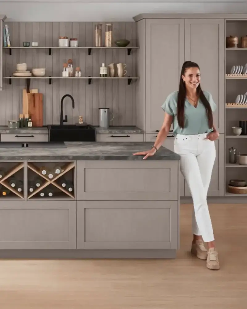 Frau mit langen dunklen Haaren, die ein grünes Oberteil, weiße Hose und bequeme Schuhe trägt und lächelnd in einer modernen, hellgrauen Küche mit Kücheninsel und Weinregal steht.