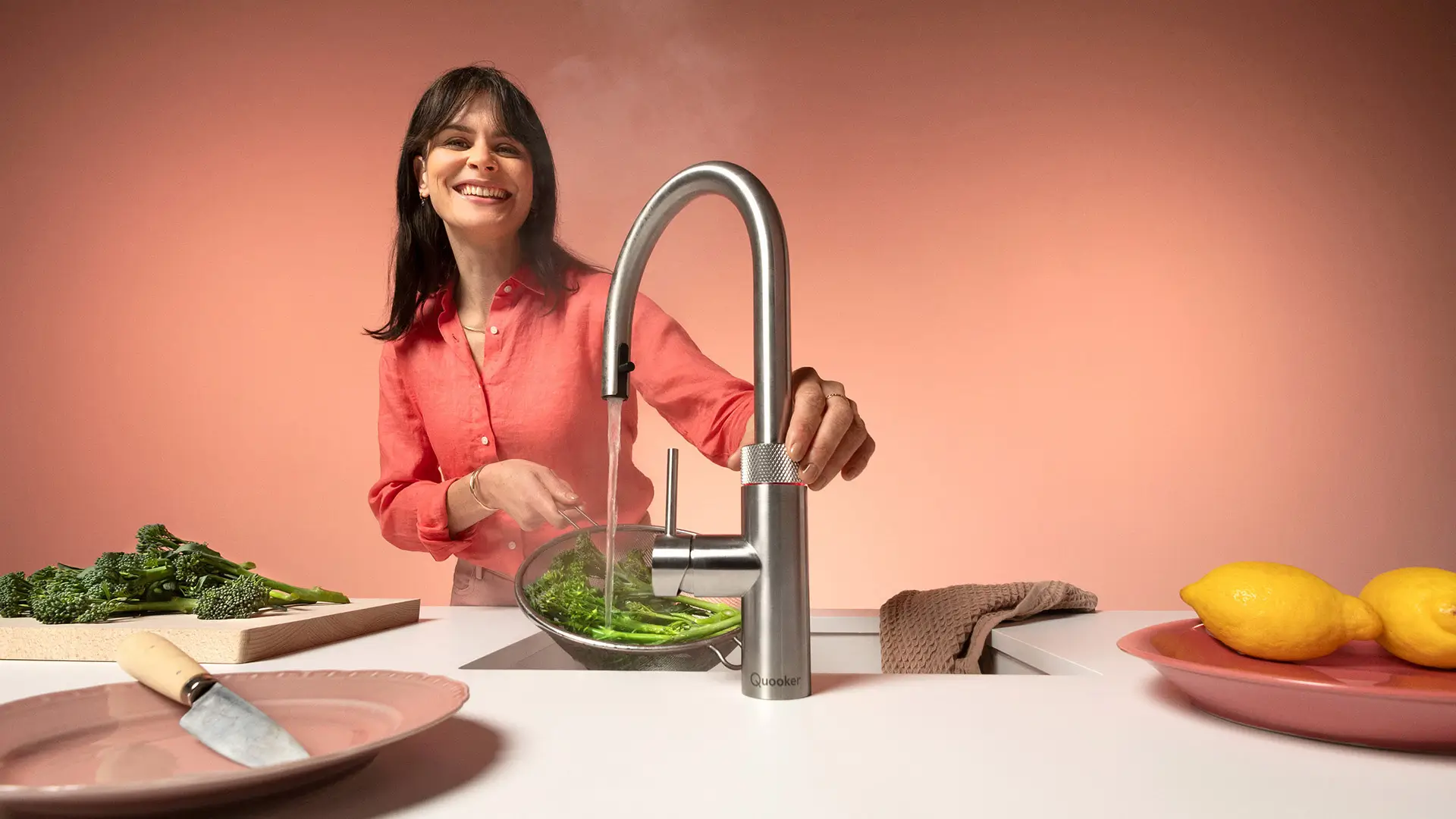 Frau in roter Bluse lächelt beim Waschen von grünem Gemüse in einem Sieb unter einem modernen Edelstahlschrank mit laufendem Wasserhahn in einer hellen Küche mit rosa Hintergrund.