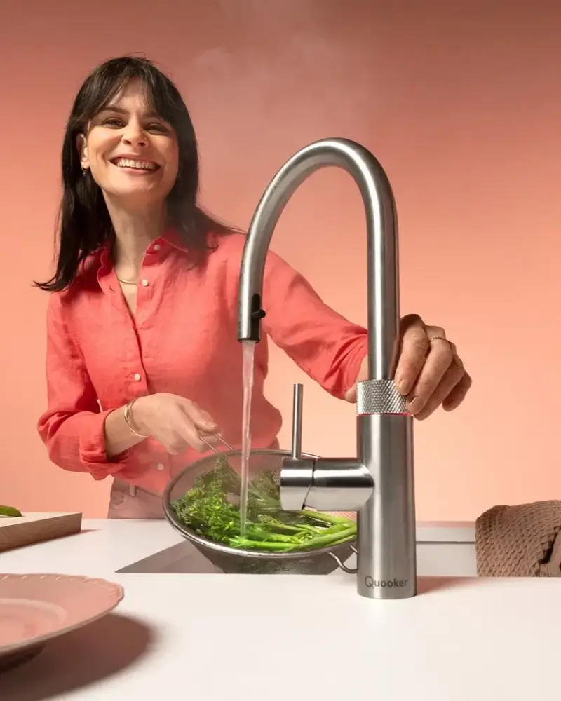 Frau in roter Bluse lächelt beim Waschen von grünem Gemüse in einem Sieb unter einem modernen Edelstahlschrank mit laufendem Wasserhahn in einer hellen Küche mit rosa Hintergrund.