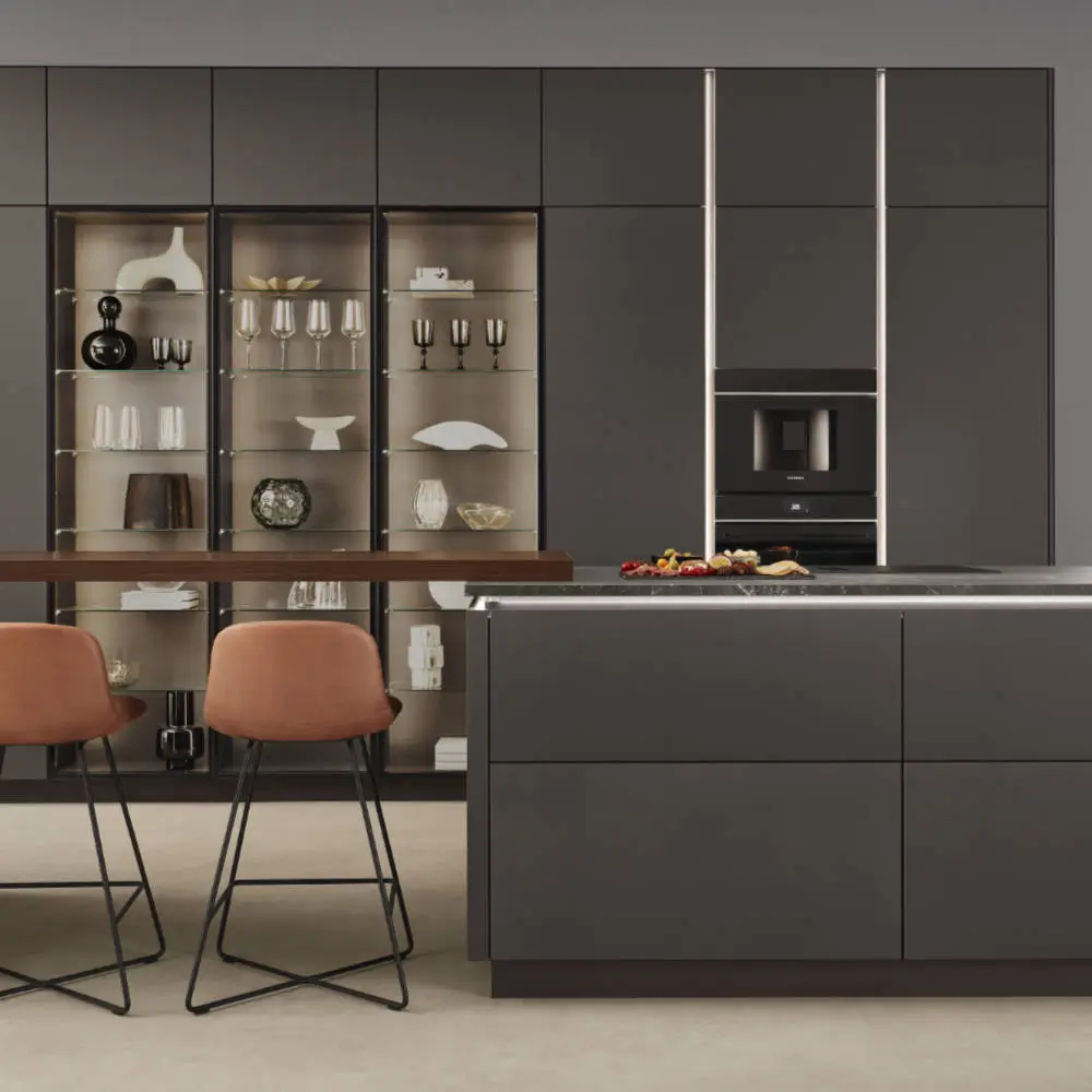 Moderne Küche mit dunkelgrauen Schränken und Insel, integriertem Backofen, Glasschrank mit Dekorationen und zwei braunen Barhockern mit schwarzen Metallgestellen.